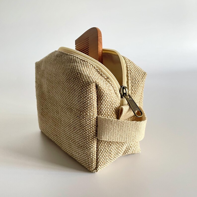 May include: A small, beige canvas toiletry bag with a zipper and a handle. A wooden comb is partially visible inside the open bag. The bag has a textured appearance and a neutral color palette.