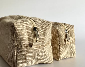 Op de afbeelding: Twee beige canvas etuis met ritssluiting. De etuis zijn rechthoekig met een geweven textuur. De ritsen zijn bronskleurig. De etuis zijn waarschijnlijk bedoeld voor het opbergen van toiletartikelen of andere kleine spullen.