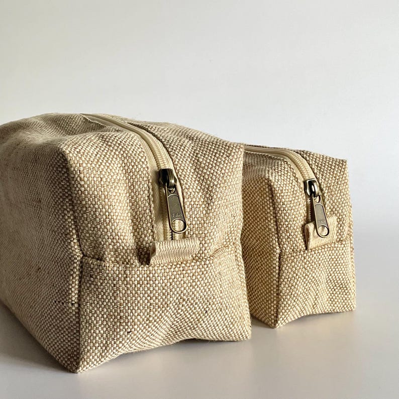 May include: Two beige canvas zippered pouches. The pouches are rectangular with a textured weave. The zippers are a bronze color. The pouches are likely for storing toiletries or other small items.