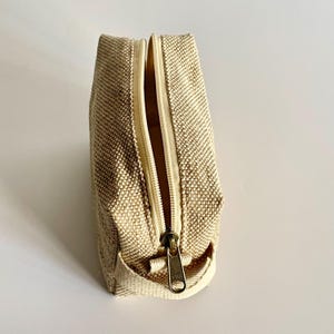 May include: A beige canvas cosmetic bag with a brass zipper. The bag is rectangular with a zipper that runs along the top. The zipper is partially open, revealing the interior. The bag is made of a woven fabric.