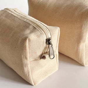 May include: Two beige canvas zippered pouches of different sizes. The smaller pouch is in the foreground, with a visible zipper and a YKK zipper pull. The larger pouch is in the background. Both pouches are made of a textured fabric.