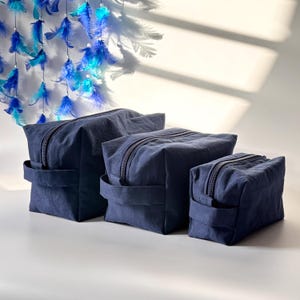 May include: Three navy blue zippered cosmetic bags of varying sizes are arranged in a row. Each bag has a rectangular shape with a handle on one side. A blue and white feather mobile hangs in the background.