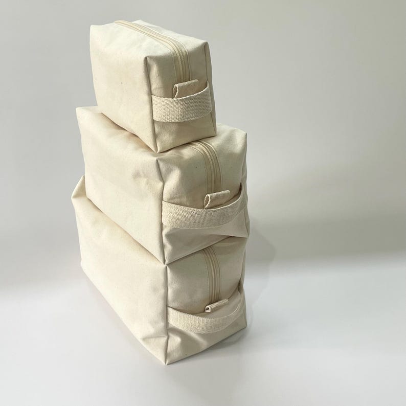 Op de afbeelding: Drie gestapelde, off-white canvas ritszakjes. Elk zakje heeft aan &eacute;&eacute;n kant een bijpassende canvas handgreep. De ritsen zijn lichtbeige van kleur. De zakjes zijn rechthoekig en lijken leeg te zijn.