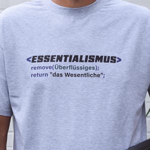 May include: A light grey crew neck t-shirt featuring dark blue text that reads <ESSENTIALISMUS> remove(Überflüssiges); return "das Wesentliche"; printed on the chest. The shirt has short sleeves and a standard fit, made from a heathered fabric.