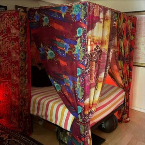 May include: A four-poster bed frame draped with sheer, multi-colored fabric panels featuring floral and abstract patterns in shades of red, yellow, and blue. The bed is covered with a striped mattress cover in white, pink, and orange hues.