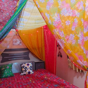 May include: A bed with a canopy made of colorful patterned fabrics. The bedspread is red with floral embroidery. Pillows in green and patterned designs are on the bed. The canopy is made of yellow, red, and patterned fabrics.