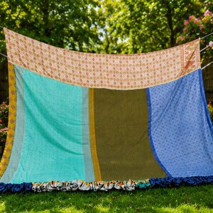 May include: A rectangular fabric throw featuring a patchwork design with a horizontal top panel in cream and orange, followed by vertical sections in turquoise, olive green, and blue with intricate patterns, finished with a dark blue ruffled edge.