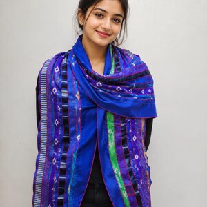 May include: A vibrant blue scarf featuring a multi-patterned design with vertical stripes, geometric diamond shapes, and accents of green and purple. The scarf has a smooth texture and finished edges, draped over the shoulders with a pointed hemline.