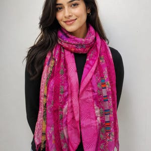 May include: A long, rectangular scarf featuring a patchwork design of various pink fabric strips. The scarf displays a mix of textures and patterns, including floral motifs, palm leaf prints, and striped sections in shades of magenta and fuchsia.