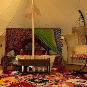 May include: A bell tent interior featuring a bed draped with red and green patterned fabrics, a central wooden console table, a woven pendant light, and a macrame hanging chair with a cream cushion, all set on a vibrant, multi-colored rug.