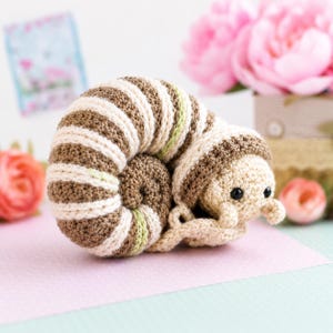 Puede incluir: Un juguete de caracol tejido a ganchillo con una concha en espiral de rayas alternas marrones y crema, acentuadas por finas líneas de color verde claro. El caracol tiene una cabeza de color crema con dos pequeñas antenas y dos ojos de cuentas negras.