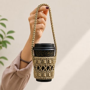 May include: A beige macrame coffee cup holder with a matching strap, holding a black paper coffee cup. The cup has a black lid and a gold pattern. The holder is made of woven cord.