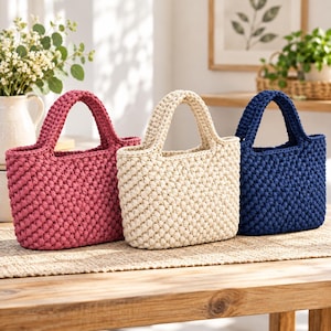 May include: Three small handbags crafted from thick, textured yarn in solid colors. The bag on the left is deep pink, the center bag is cream, and the bag on the right is navy blue. Each bag features a rounded handle and a dense, knotted stitch pattern.
