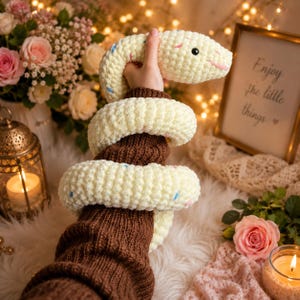 May include: A cream-coloured crocheted snake plush toy with small blue and pink speckles, featuring a black eye and a stitched pink mouth. The plush is coiled in a spiral shape, showcasing a soft, textured yarn construction throughout its body.