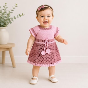 May include: A crochet dress featuring a light pink bodice and a darker mauve skirt with a scalloped hem. The dress includes a drawstring waist with two small crocheted flower accents. A matching mauve headband with a small bow is worn.
