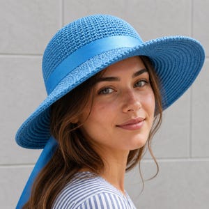 May include: A light blue sun hat with a wide brim and a matching ribbon band. The hat has a woven texture and a rounded crown. The ribbon hangs down the side. The hat is worn by a person wearing a blue and white striped shirt.