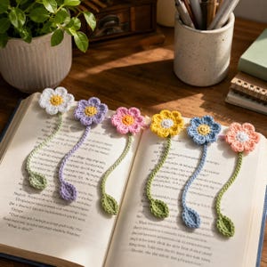 May include: A collection of six handmade crochet flower bookmarks in various colors, including white, purple, pink, yellow, blue, and peach. Each bookmark features a flower head, a stem, and a leaf, resting on the pages of an open book.