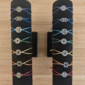 May include: Two black cylindrical displays holding fourteen cord bracelets. Each bracelet features a unique silver-toned metal charm with geometric symbols. The cords are arranged in a spectrum of colors including purple, blue, teal, green, yellow, orange, and red.