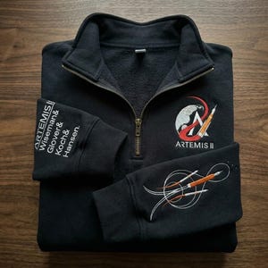 Puede incluir: Sudadera negra con cremallera de cuarto de cuello y diseño bordado de la misión Artemis II. Presenta el logotipo de Artemis II en el pecho y detalles de la misión en la manga izquierda. La manga derecha tiene un diseño de cohete estilizado.