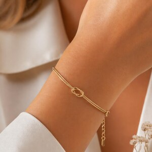 May include: A delicate gold bracelet featuring a knot design. The bracelet has a double-strand chain and an adjustable clasp. The knot is the focal point, adding a touch of elegance to the jewelry piece. The bracelet is shown on a person's arm.