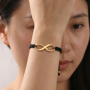 May include: A gold-colored infinity symbol bracelet with the names "Kevin" and "Karen" inscribed. The bracelet features a black woven band and a sliding knot closure for adjustable sizing. A personalized jewelry item.