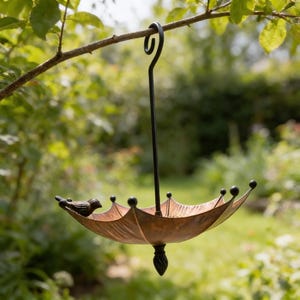 May include: A decorative bird feeder shaped like an umbrella, suspended from a tree branch. The feeder has a bronze-colored bowl with a small bird perched on the edge. The feeder is made of metal and has a black hanging hook.