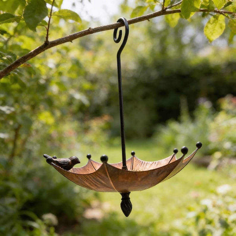 May include: A decorative bird feeder shaped like an umbrella, suspended from a tree branch. The feeder has a bronze-colored bowl with a small bird perched on the edge. The feeder is made of metal and has a black hanging hook.