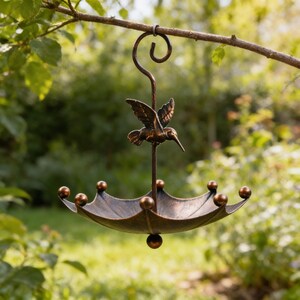 May include: A bronze-colored metal bird feeder shaped like an umbrella, suspended from a tree branch. The feeder features a hummingbird sculpture and decorative ball accents around the rim. The feeder is designed to attract birds.