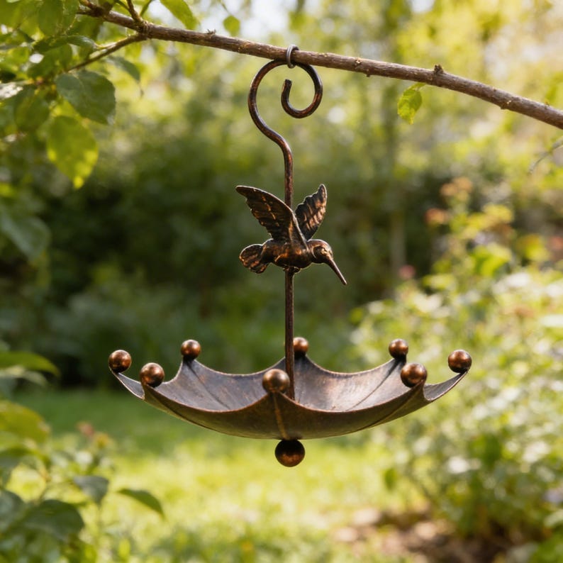May include: A bronze-colored metal bird feeder shaped like an umbrella, suspended from a tree branch. The feeder features a hummingbird sculpture and decorative ball accents around the rim. The feeder is designed to attract birds.