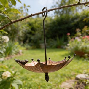 May include: A bronze-colored bird feeder shaped like an umbrella, hanging from a tree branch. The feeder has a small bird perched on the edge. The feeder is filled with birdseed and has decorative black ball accents.