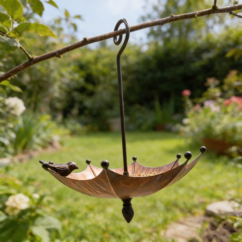 May include: A bronze-colored bird feeder shaped like an umbrella, hanging from a tree branch. The feeder has a small bird perched on the edge. The feeder is filled with birdseed and has decorative black ball accents.