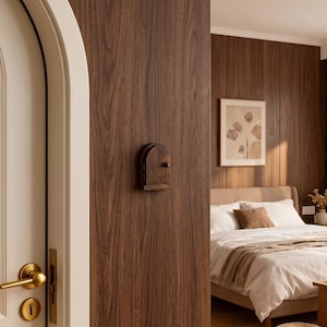 May include: A small, arched, dark brown wooden decorative door mounted on a vertical wood-paneled wall. The miniature door features a rounded top, a flat base, and a single small round knob centered on the right side of the wooden paneling.