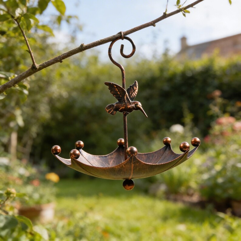 May include: A bronze-colored metal bird feeder shaped like an umbrella, suspended from a tree branch. The feeder features a hummingbird figure at the top and decorative spheres around the edge. The background shows a blurred garden scene.