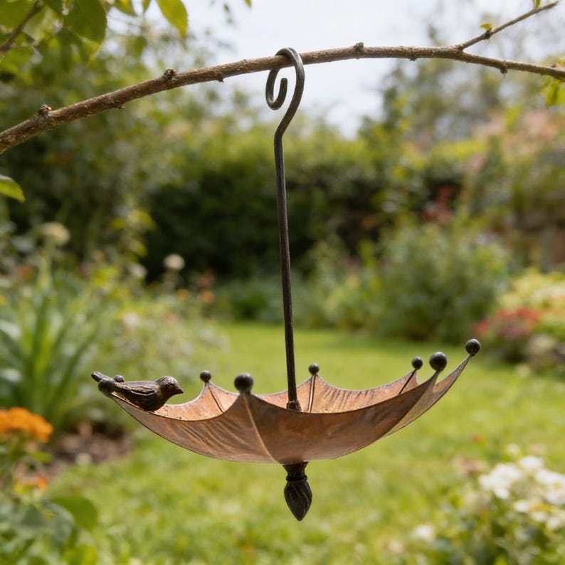 May include: A bronze-colored bird feeder shaped like an umbrella, suspended from a tree branch. The feeder has a decorative bird perched on the edge and is designed to attract birds to a garden setting.