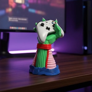 Op de afbeelding: Een kleurrijke gamecontrollerstandaard in de vorm van een hand die een witte controller vasthoudt. De standaard heeft groene, rode, roze en blauwe accenten. Hij staat op een donkerbruin bureau, met een computermonitor en een gameconsole op de achtergrond.