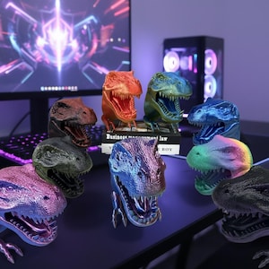 Könnte beinhalten: Eine Sammlung farbenfroher Tyrannosaurus Rex-Kopfskulpturen. Die Dinosaurierköpfe sind in verschiedenen Farben erhältlich, darunter Rot, Blau, Grün und Lila. Die Köpfe werden auf einer dunklen Oberfläche mit einem Computer und einer Tastatur im Hintergrund ausgestellt.
