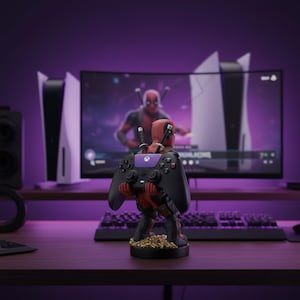 Może przedstawiać: Uchwyt na kontroler w stylu Deadpool w konfiguracji do gier. Uchwyt, przedstawiający postać w czerwono-czarnym kostiumie, trzyma czarny kontroler z fioletowymi akcentami. Tło obejmuje monitor wyświetlający obraz Deadpoola i konsole do gier.
