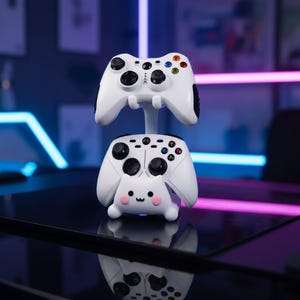以下が含まれることがあります： キュートな漫画デザインの白いゲームコントローラー用スタンド。スタンドは2つの白いゲームコントローラーを保持しており、1つは上、もう1つは下にあります。下のコントローラーには、ピンクの頬の顔があります。