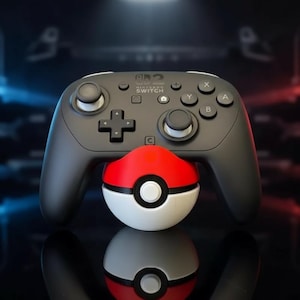 Puede incluir: Un mando Nintendo Switch Pro negro con joysticks y botones blancos, apoyado en una Pokeball roja, blanca y negra. El mando tiene el logotipo de Nintendo Switch.