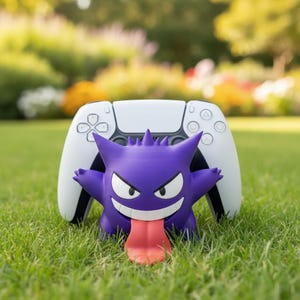 Può includere: Una figura viola di Gengar con lingua rossa, occhi bianchi e un sorriso minaccioso, davanti a un controller bianco di PlayStation 5. La figura è su erba verde, con uno sfondo sfocato di alberi e fogliame.