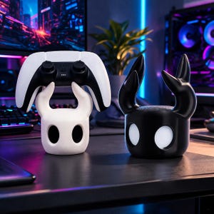 May include: A white and black gaming controller sits on a white, stylized holder. Next to it, a black holder with glowing white eyes and horns. The items are on a dark surface, with a computer and plant in the background.
