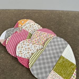Könnte beinhalten: Ein Stapel herzförmiger Stoffuntersetzer mit verschiedenen Mustern. Die Untersetzer weisen eine Mischung aus Farben auf, darunter Rosa, Grau, Grün und Orange. Jeder Untersetzer hat ein einzigartiges Patchwork-Design.