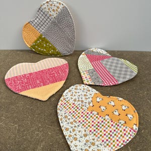 Könnte beinhalten: Vier herzförmige Stoffuntersetzer mit Patchwork-Designs. Jeder Untersetzer hat eine einzigartige Kombination aus Mustern und Farben, darunter Rosa, Orange, Grün und Grau. Die Untersetzer sind wahrscheinlich handgefertigt.