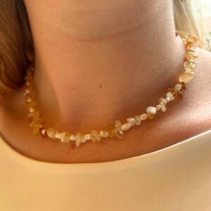 Peut inclure: Un collier composé d'un mélange de petites perles blanches irrégulières et de fragments de pierres précieuses jaune-orange translucides. Les perles sont enfilées en alternance, créant un aspect texturé autour du cou. Une boucle d'oreille assortie est visible.