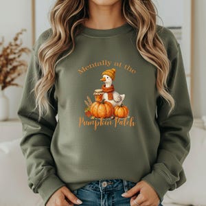 May include: An olive green crewneck sweatshirt featuring a graphic of a white goose wearing an orange knit hat and scarf, sitting next to two orange pumpkins and a jar of honey, with the text Mentally at the Pumpkin Patch printed in orange.