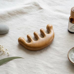 May include: A wooden scalp massager with a curved handle and rounded tines. A small amber glass bottle with a white label and a wooden cap is next to the massager. A small bowl of white crystals and a gray stone are also in the image.