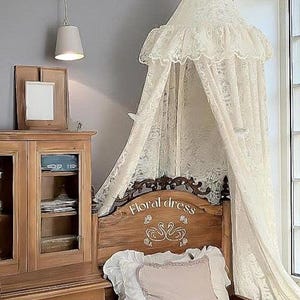 Puede incluir: Una escena de dormitorio con una cama de madera con un cabecero que dice "Floral dress". La cama está adornada con ropa de cama blanca y una almohada rosa. Un dosel de encaje color crema cuelga sobre la cama. También se ven un armario de madera y una lámpara colgante.