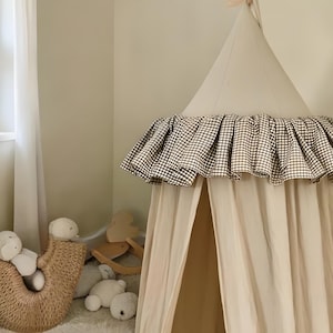 Puede incluir: Una tienda de campaña de juego de estilo tipi beige y blanco con un ribete de volantes a cuadros. La tienda está en la habitación de un niño, con animales de peluche y una cesta de mimbre cerca. Se ve una ventana con cortinas blancas.