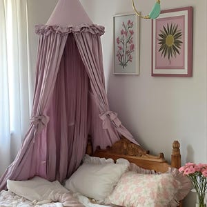May include: A light purple canopy bed with a ruffled edge, tied with bows. The bed is adorned with white pillows and a floral patterned pillow. Two framed floral prints hang on the wall, along with a gold and turquoise wall hanging.