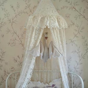 May include: A cream-colored lace bed canopy featuring a conical top with a ruffled edge and long, flowing panels that drape downward. Suspended from the center of the canopy is a plush swan figure with outstretched wings and a small floral crown.
